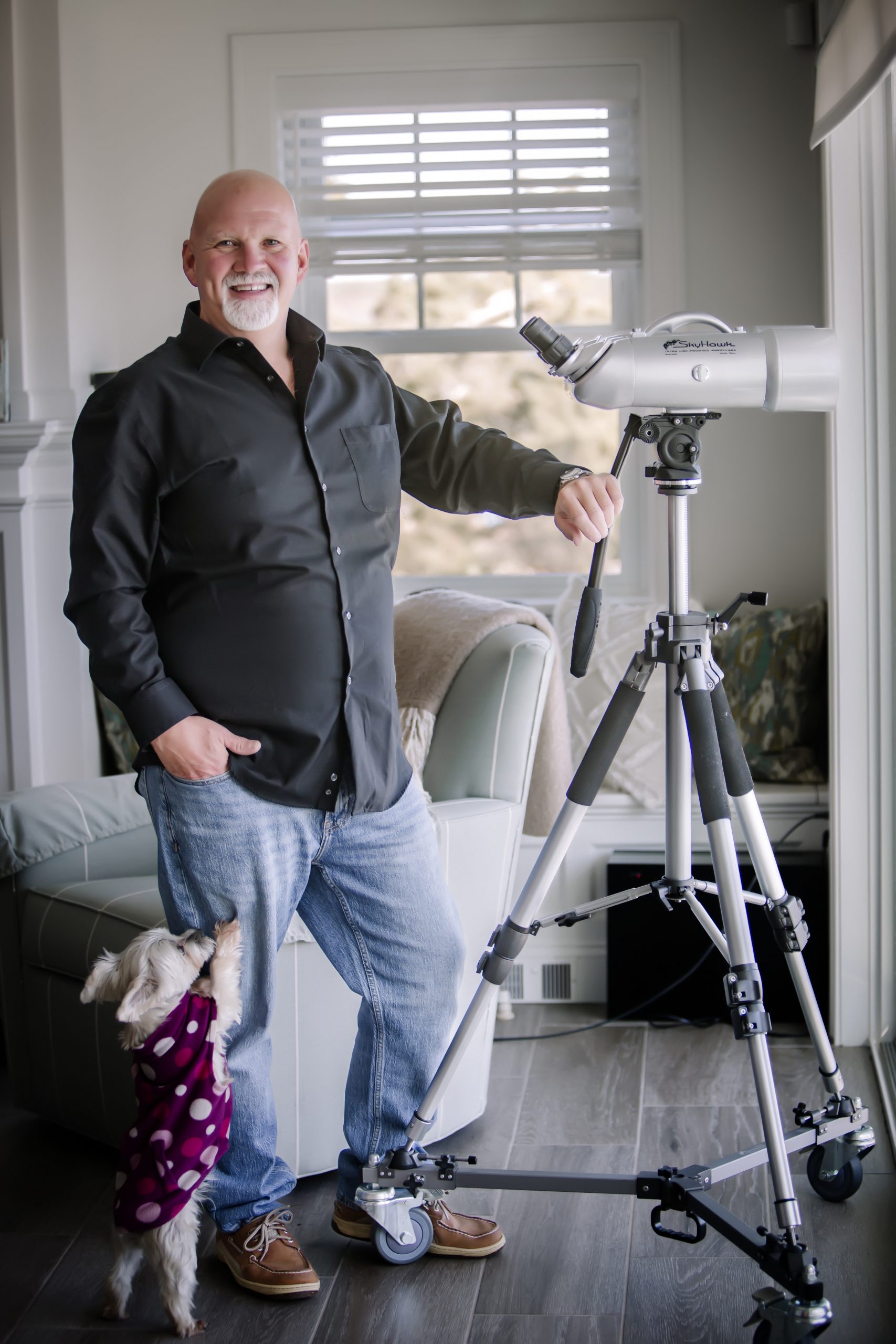 man standing in living room next to a telescope, small dog jumping on his leg