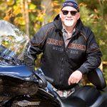 Man standing behind harley davidson wearing dew rag, leather jacket & sunglasses