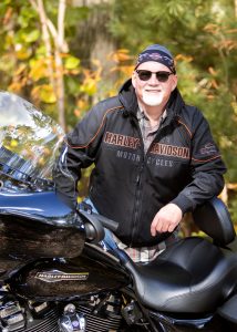 Man standing behind harley davidson wearing dew rag, leather jacket & sunglasses