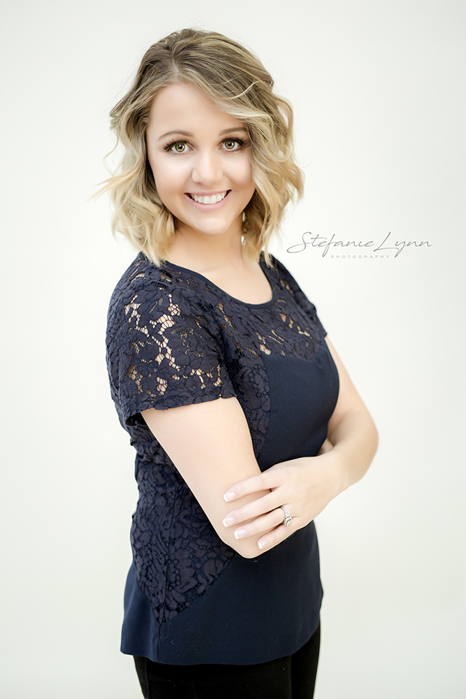 3/4 Headshot of woman in blue top standing infant of cream background, smiling withers crossed